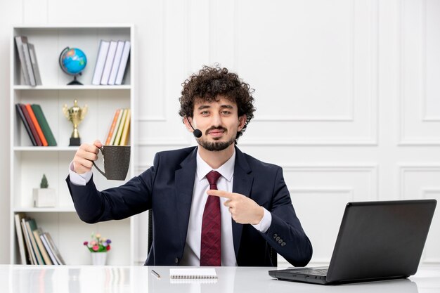 Servicio al cliente apuesto joven en traje de oficina con computadora portátil y auriculares sonriendo con taza de café