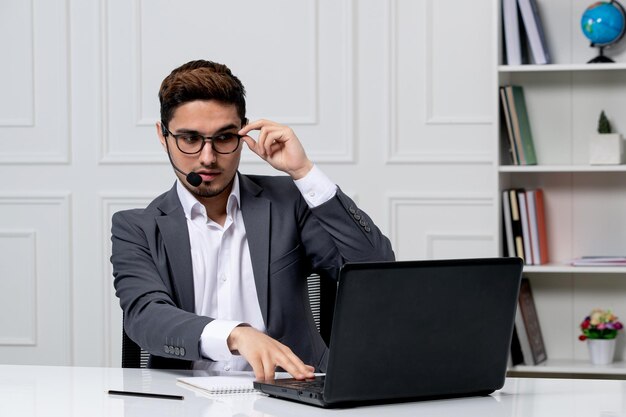 Servicio al cliente bastante caballero con computadora en traje de oficina gris tocando el borde de las gafas