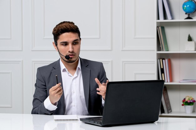 Servicio al cliente joven chico lindo en traje de oficina gris con computadora sosteniendo un lápiz mientras está de guardia