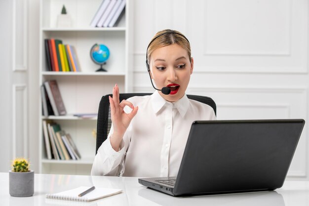 Servicio al cliente linda chica hermosa con camisa blanca con computadora y auriculares que muestran un gesto correcto