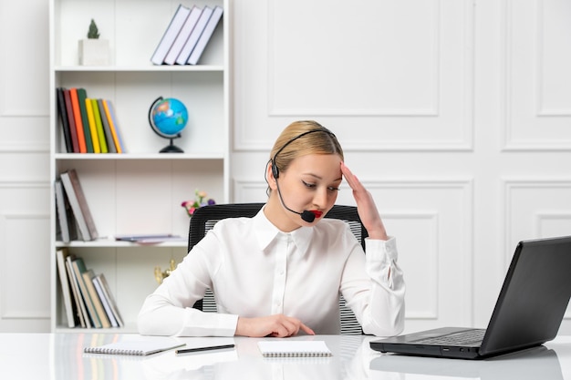 Servicio al cliente linda chica rubia con camisa blanca con computadora portátil y auriculares cansados sosteniendo la cabeza