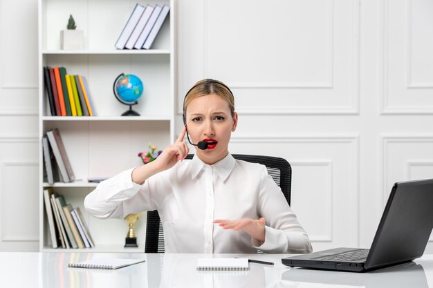 Servicio al cliente linda chica rubia con camisa blanca con computadora portátil y auriculares irritada por un cliente