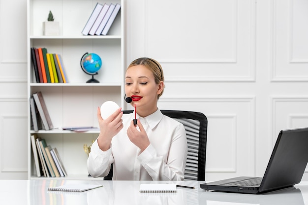 Servicio al cliente linda chica rubia con camisa blanca con computadora portátil y auriculares poniéndose lápiz labial