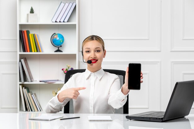 Servicio al cliente linda chica rubia con camisa blanca con computadora portátil y auriculares sonriendo sosteniendo el teléfono