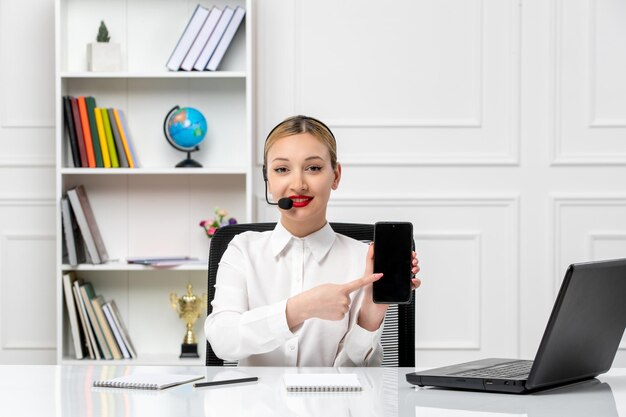 Servicio al cliente linda chica rubia con camisa blanca con computadora portátil y auriculares sonriendo con teléfono