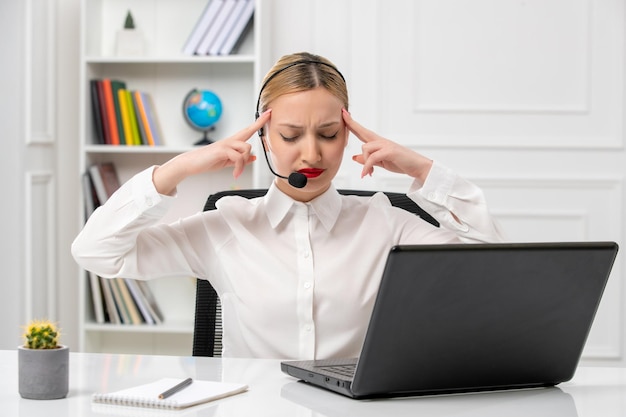 Servicio al cliente linda chica rubia con camisa blanca con computadora portátil y auriculares tocando las sienes