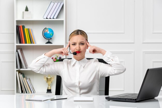 Servicio al cliente linda chica rubia con camisa blanca con computadora portátil y auriculares tocando las sienes