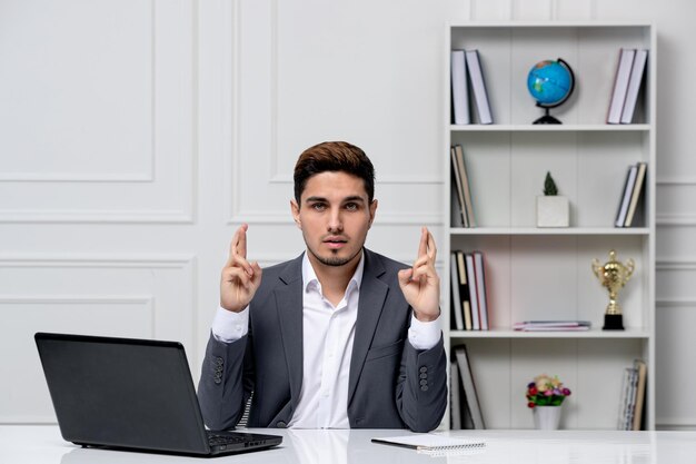 Servicio al cliente lindo chico guapo en traje de oficina con computadora cruzando los dedos y esperando