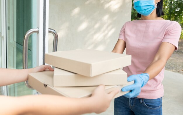 Servicio de entrega en camiseta, con máscara protectora y guantes dando orden de comida, sosteniendo tres cajas de pizza en el frente de la casa, mujer aceptando entrega de cajas del repartidor durante el brote de COVID-19.