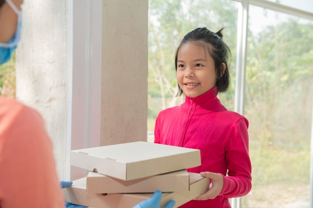 Servicio de entrega en camiseta, con máscara protectora y guantes dando orden de comida, sosteniendo tres cajas de pizza en el frente de la casa, mujer aceptando entrega de cajas del repartidor durante el brote de COVID-19.