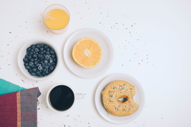 Foto gratuita servilleta; arándano; vaso de jugo; naranja a la mitad; taza de café y buñuelo sobre fondo blanco