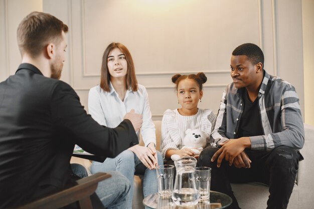 Sesión de consejería familiar a domicilio con terapeuta. Psicólogo mostrando imágenes de emociones a una niña. Padre afroamericano y madre europea.