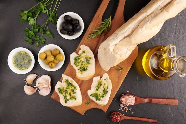 Set de snacks mediterráneos. Aceitunas, aceite, hierbas y ciabatta en rodajas sobre una tabla de madera sobre tablero de piedra pizarra negra sobre superficie oscura, vista superior. Endecha plana.