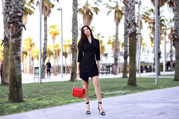 Sexy atractiva mujer segura caminando calle con palmeras, vestido negro minimalista de empresaria moderna, viajando a España.