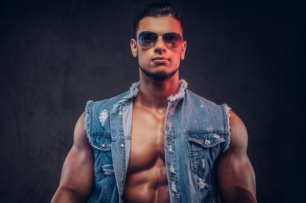 Sexy hombre desnudo de moda confiado con un cuerpo musculoso en un chaleco de mezclilla y poses de gafas de sol en un estudio. Aislado sobre fondo oscuro.