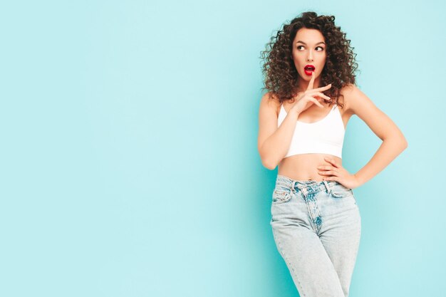 Sexy joven hermosa mujer sonriente en ropa de moda de verano hipster mujer despreocupada posando junto a la pared azul en el estudio Modelo morena positiva con peinado de rizos afro Alegre y feliz