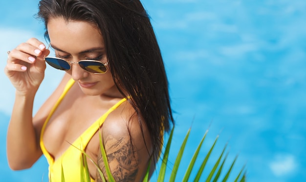 Sexy mujer morena con bronceado dorado y tatuaje, vistiendo bikini con gafas de sol, relajándose al lado de la piscina.