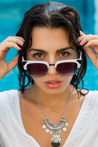 Sexy mujer morena sentada junto a la piscina con ropa de verano con estilo. Vacaciones tropicales.