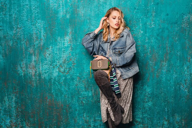 Sexy mujer rubia con estilo atractivo en jeans y chaqueta de gran tamaño caminando contra la pared verde vintage en la calle