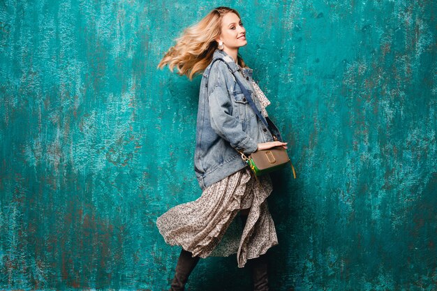 Sexy mujer rubia con estilo atractivo en jeans y chaqueta de gran tamaño caminando contra la pared verde vintage en la calle