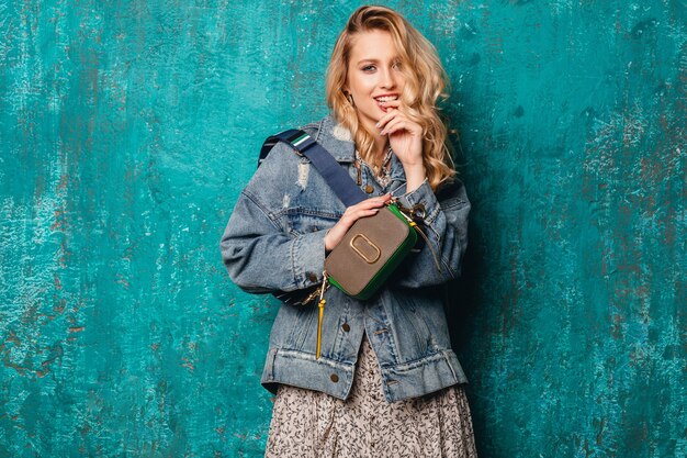 Sexy mujer rubia con estilo atractivo en jeans y chaqueta de gran tamaño caminando contra la pared verde vintage en la calle