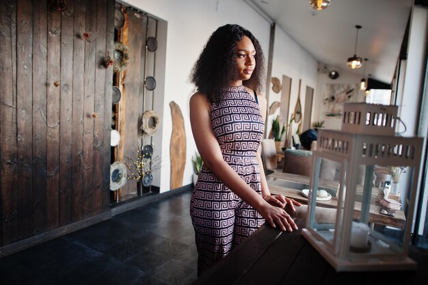 Sharming elegante joven mujer afroamericana con pelo largo y rizado con mono posando en el café interior