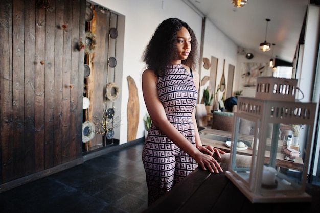 Sharming elegante joven mujer afroamericana con pelo largo y rizado con mono posando en el café interior