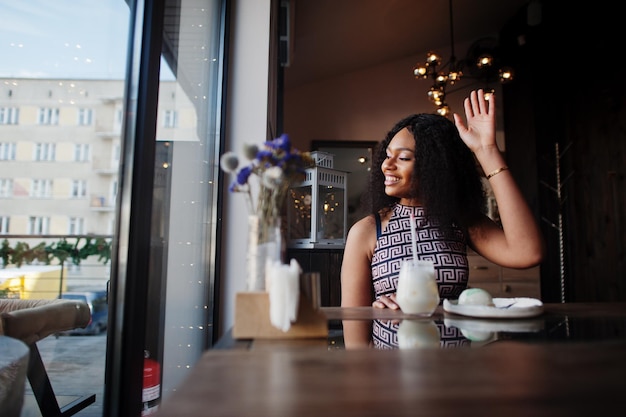 Sharming elegante joven mujer afroamericana con el pelo largo y rizado con mono sentado en el café interior y saluda a los amigos