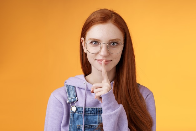 Shh caja fuerte secreta. Joven sensual misteriosa pelirroja atractiva mujer que oculta información decir callar silencio silencio por favor preparar sorpresa sostener dedos índice boca tabú, de pie fondo naranja