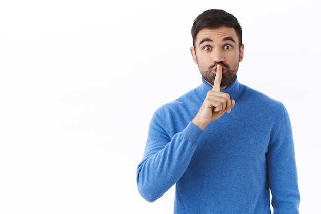 Shhh, baja la voz. Chico caucásico guapo entusiasta preparar sorpresa, pidiendo silencio, silencio con el dedo índice presionado en los labios, necesita silencio, pared blanca de pie