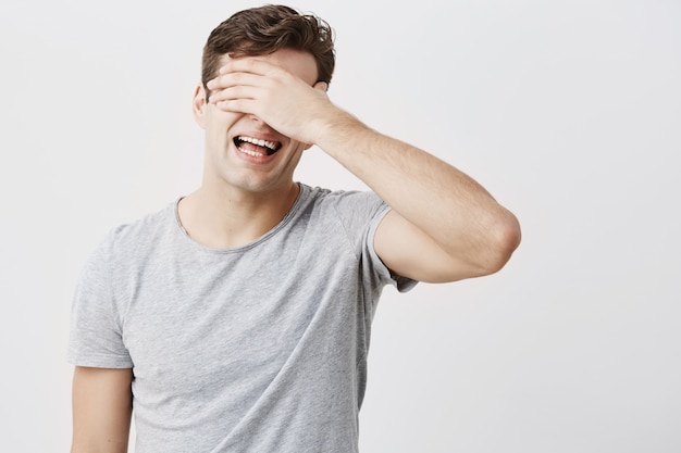 Shocked hizo hincapié en el joven emocional que ocultaba su rostro detrás de su mano, preocupado por escuchar los consejos de sus padres aislados contra el fondo de la pared del estudio en blanco. Hombre europeo cansado y molesto