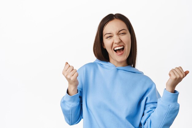 Sí, bebé, soy la ganadora Chica celebrando la victoria, logra el objetivo de éxito gritando sí y hace un gesto de bomba de puño triunfando ganando el premio en dinero de pie en una sudadera con capucha contra el fondo blanco