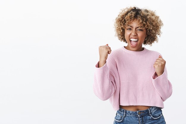 Sí, cariño, lo hicimos. Triunfando, encantada y alegre, guapa mujer afroamericana gritando de alegría y felicidad mientras ganaba apretando los puños en gesto de victoria animando sobre la pared blanca