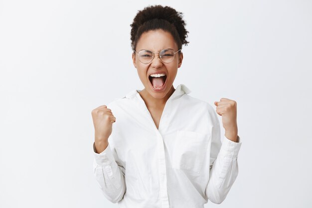 Sí chicas, lo hicimos. Retrato de triunfante atractiva mujer africana con camisa blanca y gafas, levantando los puños cerrados y gritando de asombro y felicidad, celebrando la victoria sobre la pared gris