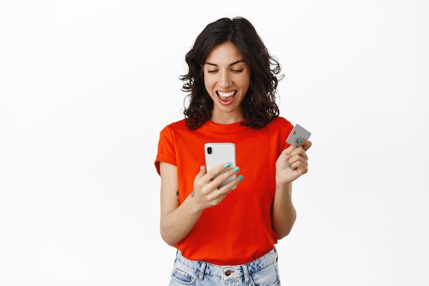 Sí, ella ganó dinero. Chica feliz grita y mira la pantalla del teléfono inteligente con cara sonriente complacida, tiene tarjeta de crédito, gana, compra con descuento, fondo blanco