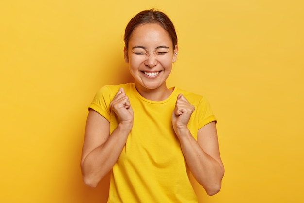 ¡sí, finalmente el éxito! la alegre chica coreana aprieta los puños con triunfo, cierra los ojos de la felicidad y la alegría, tiene una sonrisa con dientes, viste un atuendo informal, aislado en la pared amarilla, triunfa la victoria