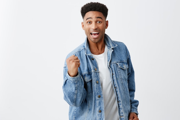 Si. Finalmente. Lo hicimos. Alegre joven atractivo hombre de piel negra con peinado afro en chaqueta de mezclilla con la mano delante de él con expresión emocionada, feliz de haber ganado la lotería.