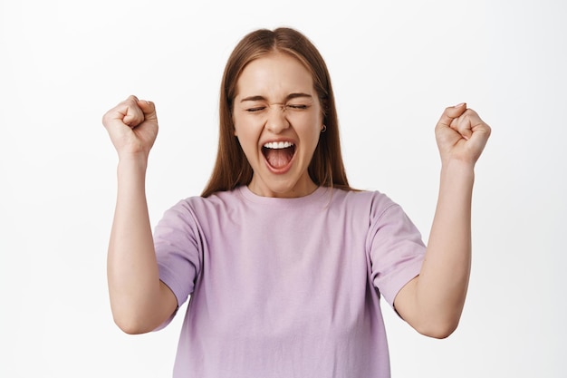 Si ganador. Una chica rubia alegre grita de felicidad y regocijo, sacudiendo los puños apretados mientras celebra, triunfa, logra la victoria de la meta, de pie contra el fondo blanco