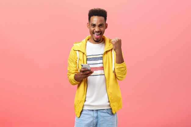 Sí, gané. Retrato de hombre afroamericano feliz emocionado y satisfecho con barba levantando el puño en gesto de victoria y triunfo regocijándose sosteniendo el teléfono inteligente ganando en el juego de teléfono en línea sobre la pared rosa