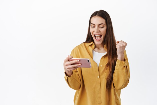 Sí, gané. Sonriente mujer feliz ganando en línea, videojuego en el teléfono inteligente, mantenga el teléfono en posición horizontal y diga sí, regocijándose y haciendo un gesto de bomba de puño para triunfar, lograr el éxito de la meta, fondo blanco