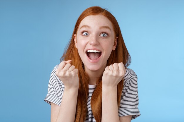 Sí, una gran noticia, genial. Atractiva chica pelirroja sorprendida animando felices puños apretados gritando sí lograr la meta ojos muy abiertos emocionados celebrando ganar lotería triunfando con alegría, fondo azul.