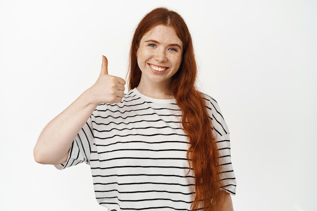 Si. Modelo de mujer pelirroja sonriente mostrando los pulgares para arriba. Chica con peinado largo rojo y pecas le gusta algo bueno, dar su aprobación, estar de acuerdo o elogiarte, de pie sobre fondo blanco.