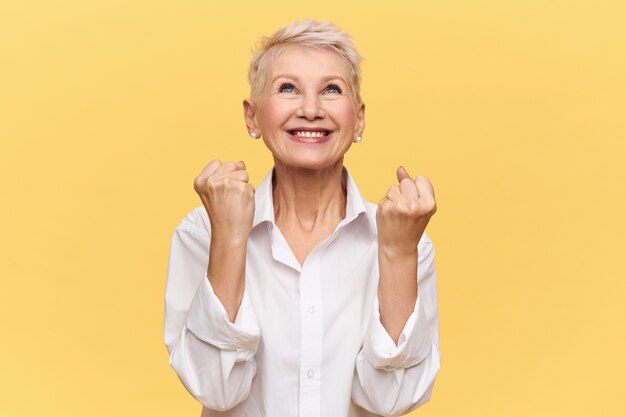 ¡Si! Mujer de mediana edad enérgica y de moda que se regocija por su éxito, alcanza metas y cumple sueños, se llena de alegría y está extasiada, cierra los puños y mira hacia arriba, agradeciendo a Dios o al Universo