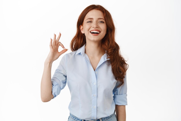 Si muy bien. Feliz mujer de negocios sonriente con el pelo rojo y rizado, asiente complacida, muestra un gesto de OK OK y ríe, aprueba y le gusta algo agradable, alaba el gran trabajo, fondo blanco