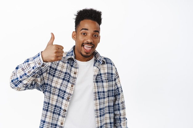 Si muy bien. Hombre negro sonriendo, mostrando el pulgar hacia arriba en aprobación, como y de acuerdo. Hombre afroamericano alaba el buen trabajo, recomienda la venta, de pie sobre fondo blanco