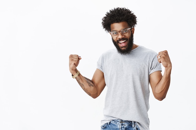 Sí, sintiendo coraje y disposición para lograr el éxito. hombre barbudo afroamericano optimista confiado y encantado que levanta los puños cerrados en celebración, triunfando siendo feliz con buen resultado