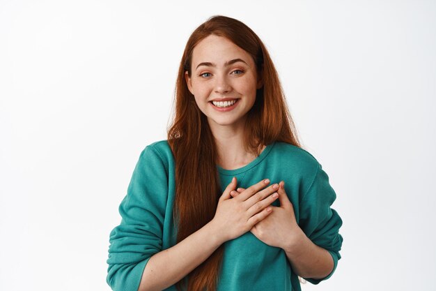 Siempre en mi corazón. Mujer joven sonriente con cabello rojo, sintiéndose conmovida y agradecida, diga gracias, sintiéndose sincera, de pie contra el fondo blanco