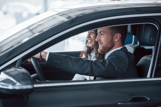 Se siente bien. Encantadora pareja exitosa probando coche nuevo en el salón del automóvil