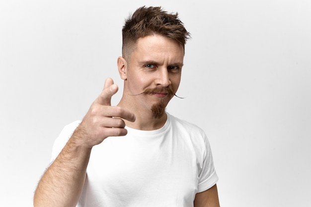 Signos, símbolos, gestos y concepto de lenguaje corporal. Vista horizontal del chico guapo joven alegre seguro con peinado de moda, bigote y barba sonriendo y apuntando con el dedo índice a la cámara