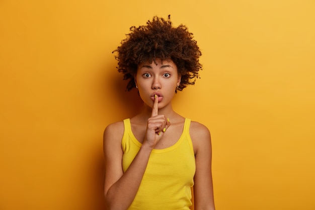 Silencio y concepto secreto. Mujer afroamericana sorprendida presiona el dedo índice contra los labios, pide silencio, dice información muy privada, usa un chaleco amarillo informal, posa, dice silencio o silencio